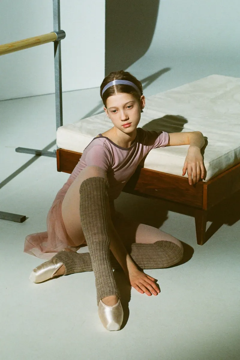 A dancer sitting on a studio floor in a pink leotard, tulle skirt, knitted leg warmers and pointe shoes