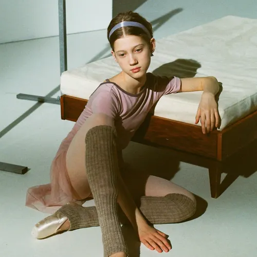A dancer in a pink leotard, tulle skirt and knitted leg warmers sitting on a studio floor