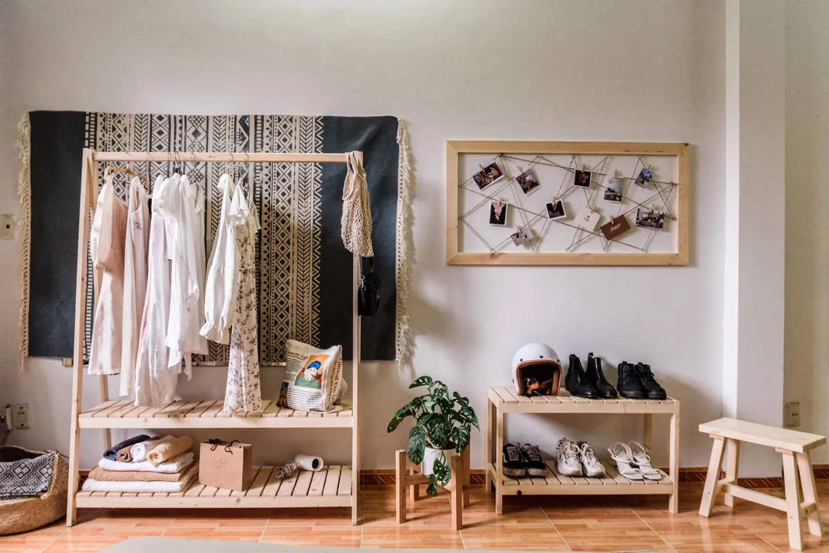 A small wooden clothes rail holding a few cream and white garments, with shoes on a low shelf and a stool beside it in a bright room