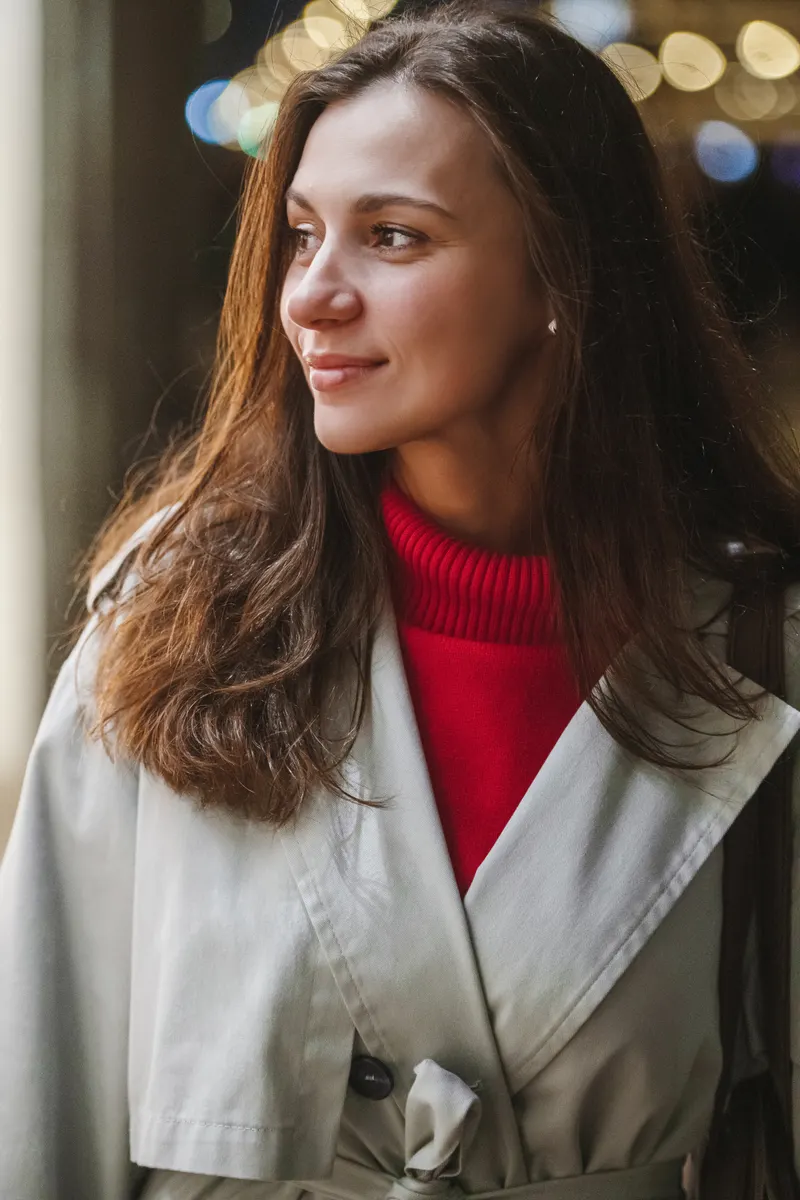 A woman with long layered brown hair falling well past her jaw, wearing a red jumper and a pale trench coat
