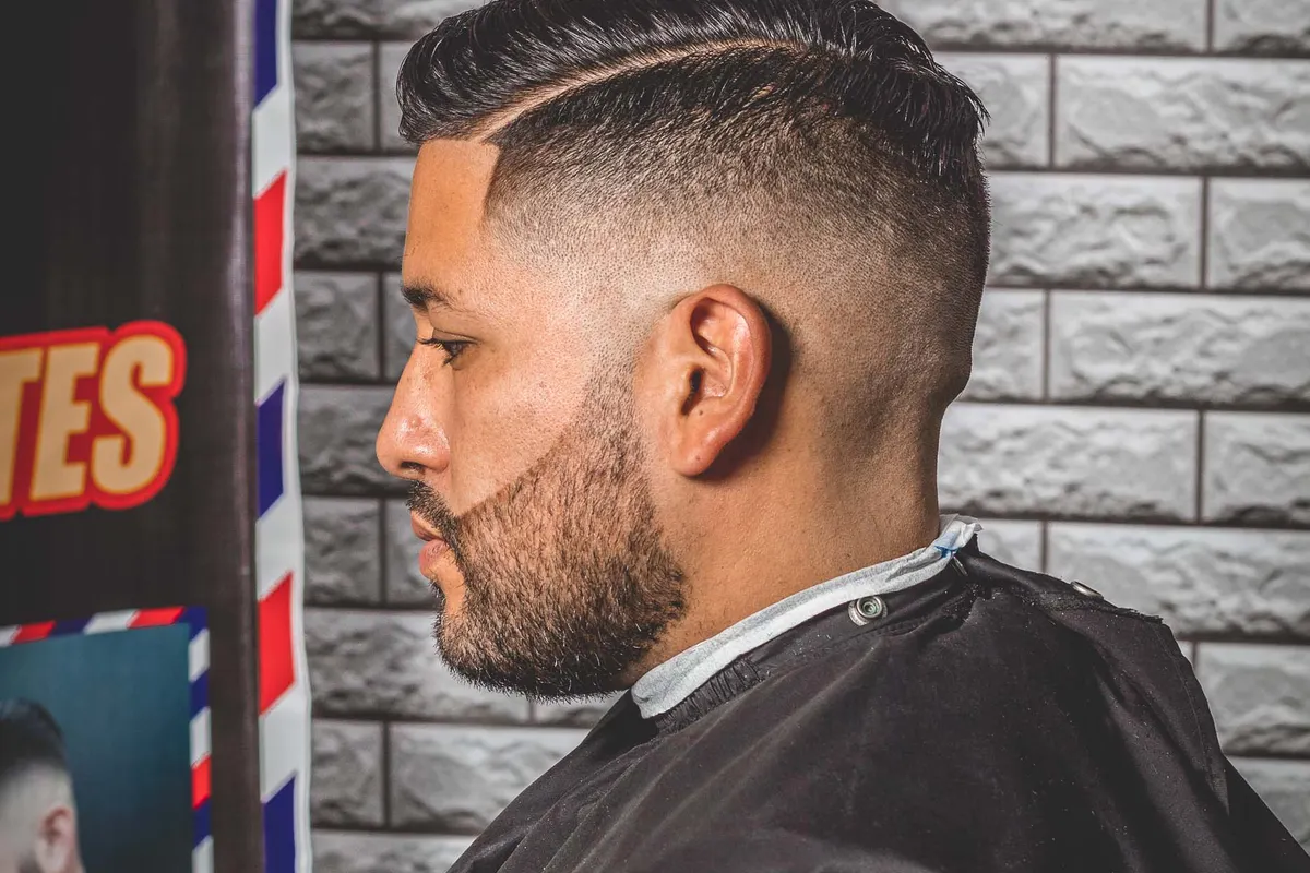 A man in a barber chair seen from the side, with a high fade at the sides and length swept up on top