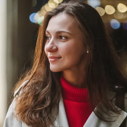 A woman with long layered brown hair wearing a red jumper and a pale trench coat