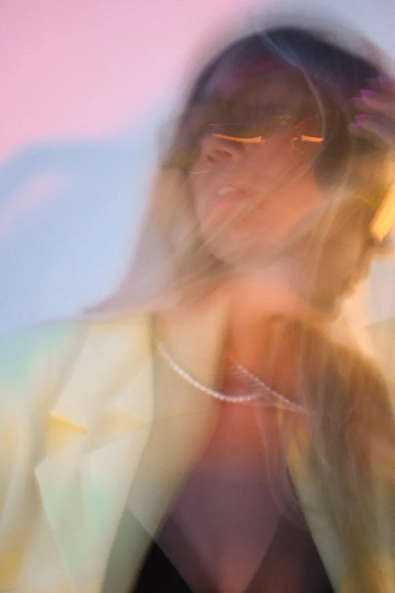 A motion-blurred portrait of a woman in sunglasses against a pastel pink and blue background