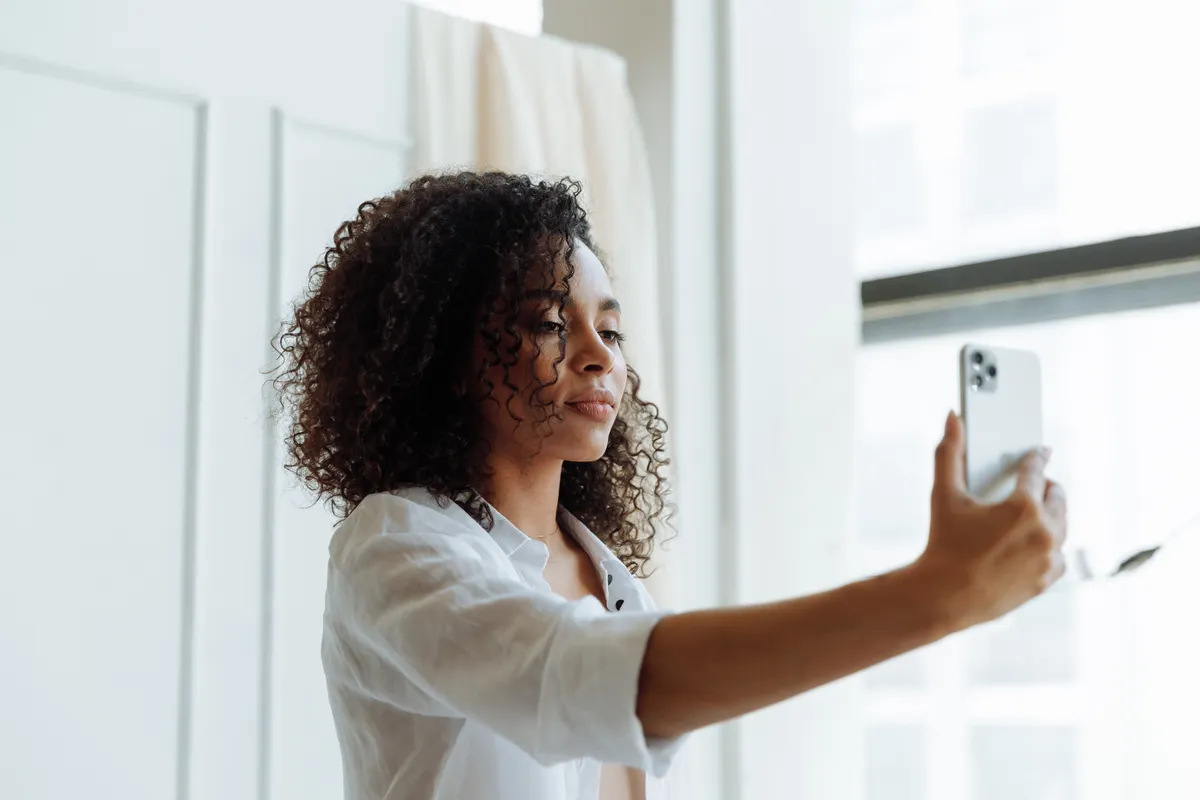 A woman with curly hair holding her phone up to take a selfie in front of a bright window