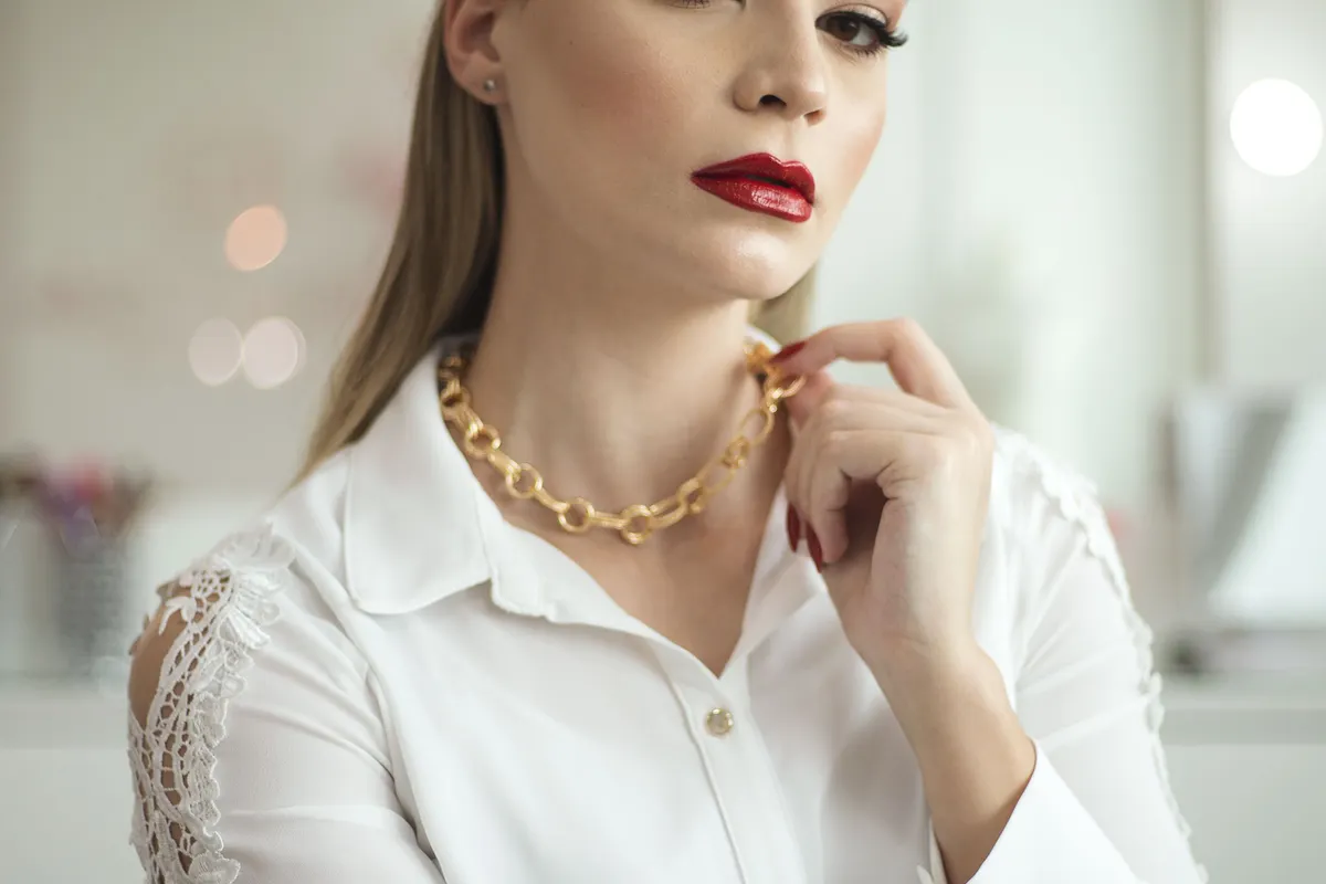 A woman wearing a dark red lip, red nail polish and a chunky gold chain necklace over a white shirt