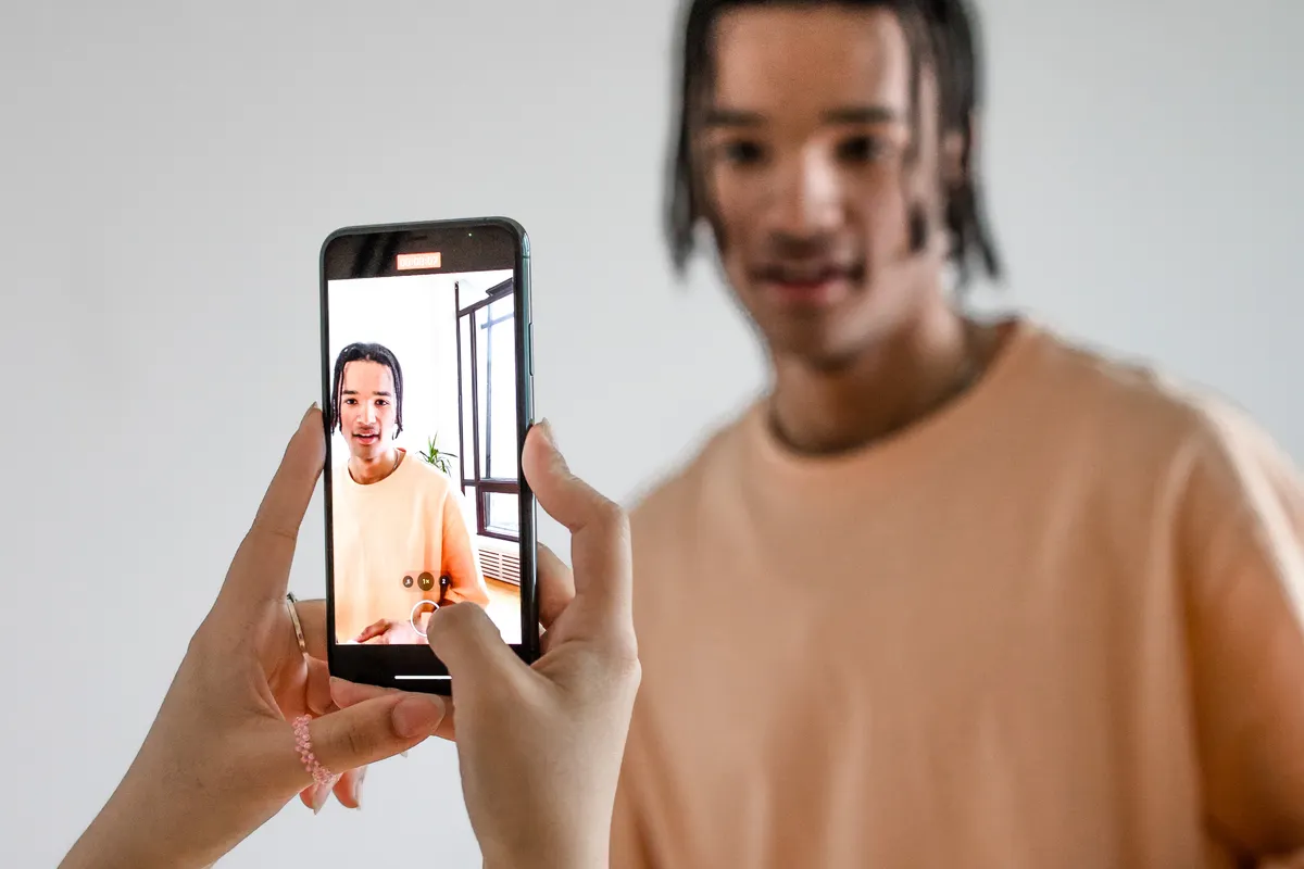 A person holding up a phone to photograph a young man in a peach T-shirt standing in front of a plain white wall