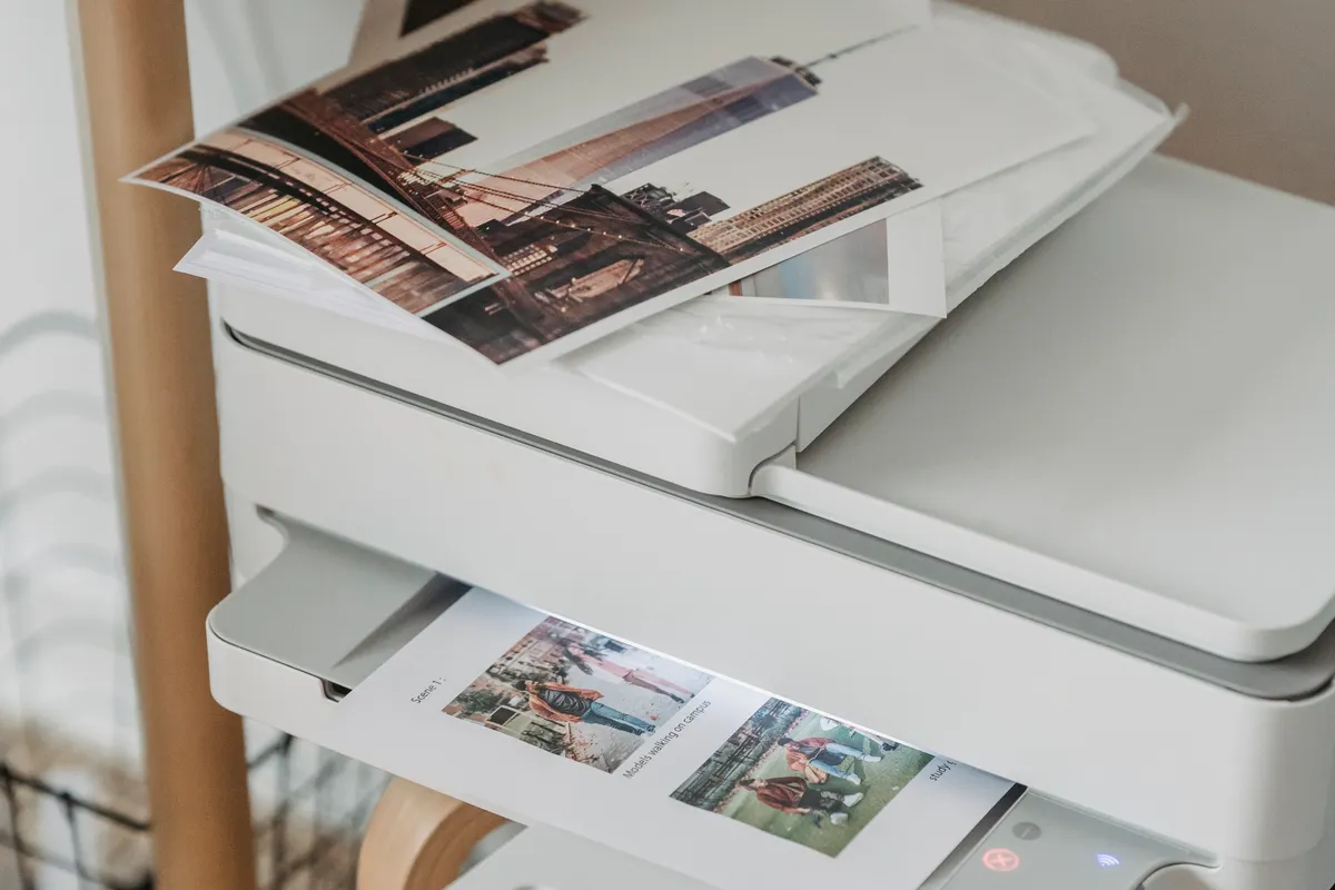 A white home printer on a wooden stand with printed photographs in its output tray and lying on the lid