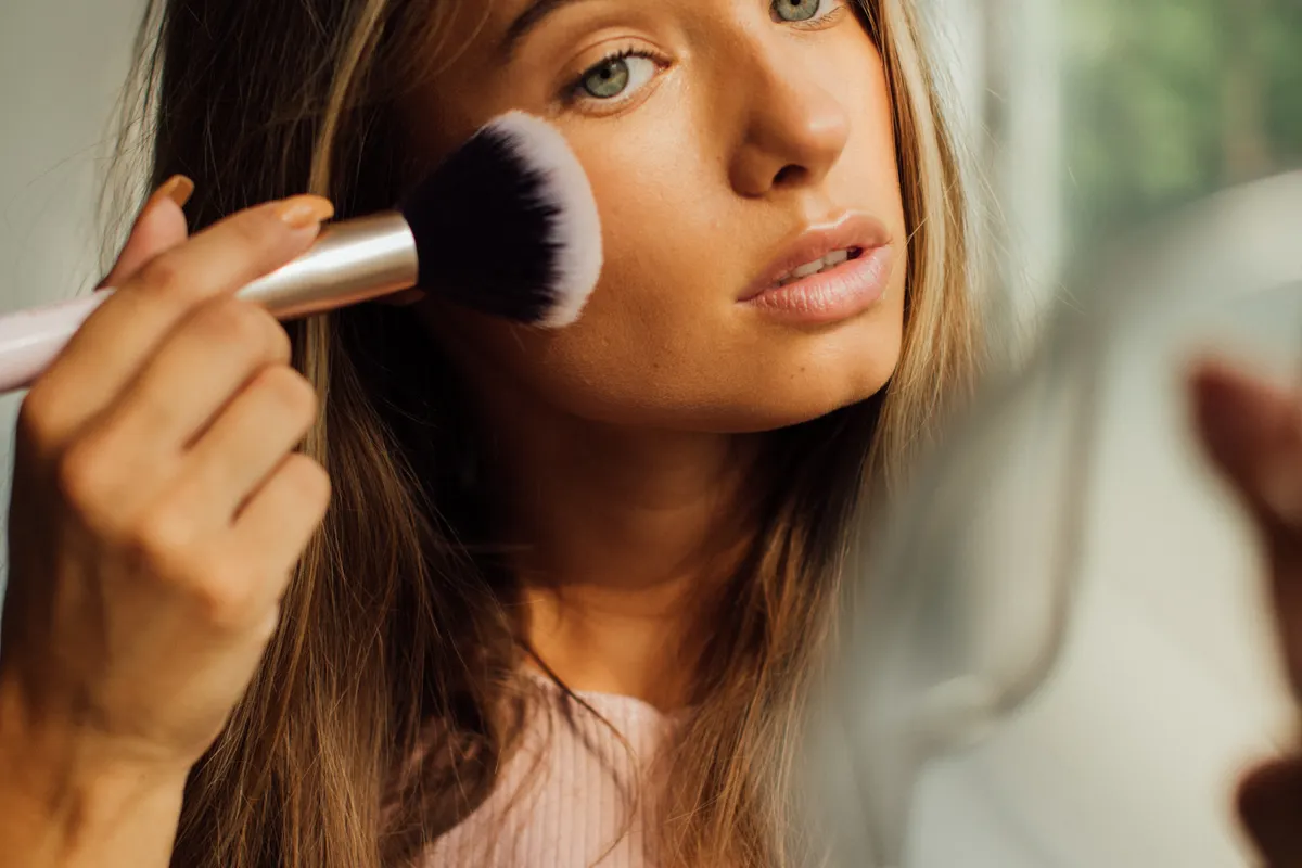 A woman by a window applying blush to her cheekbone with a large fluffy brush